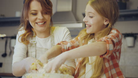 Young-beautiful-mother-teaching-her-pretty-daughter-to-cook-and-knead-a-daugh-for-cookies-on-the-table-in-the-kitchen.-Inside